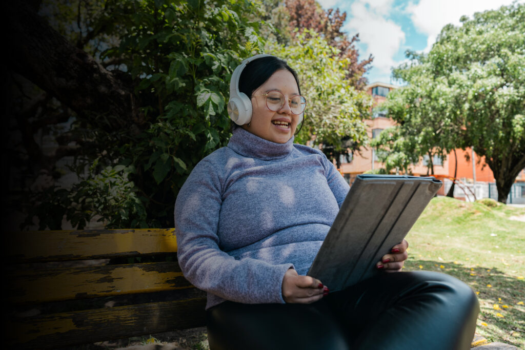 A photo of a young woman with head phones on at a park bench looking at her tablet.